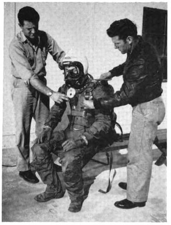 A man in a suit and helmet sitting as two standing men adjust the suit